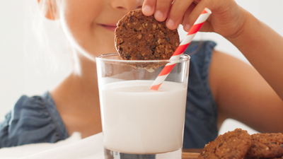 A smiling child is dipping a Real Treat Oatmeal Raisin cookie into a glass of milk.