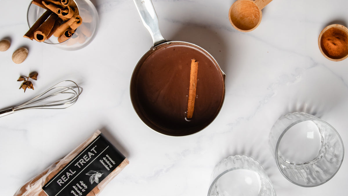 A pot of rich dark liquid chocolate on a marble counter with spices, cups, and a package of Real Treat drinking chocolate