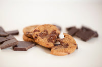 A single Real Treat Pantry Chocolate Chunk Cookie is fresh from the oven with melted chocolate and rests next to some chunks of dark chocolate. 