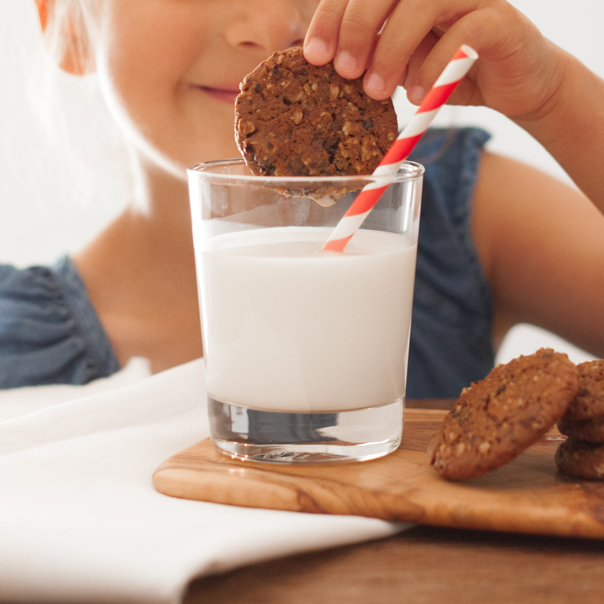 A little girl smiles and dunks a Real Treat organic oatmeal raisin cookie into a glass of milk. 