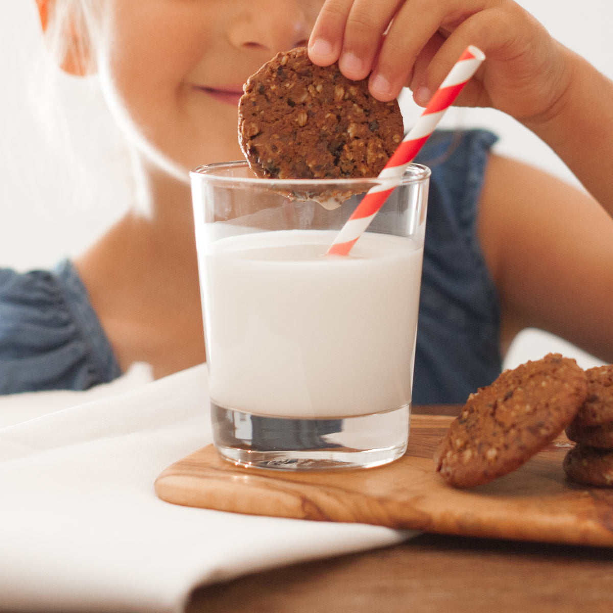 A little girl smiles and dunks a Real Treat organic oatmeal raisin cookie into a glass of milk.