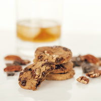 A stack of Dark Chocolate chunk with Smoked Pecans cookies sits next to a glass of bourbon and a scattering of pecans and dark chocolate.
