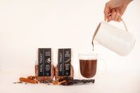 1 package of Ancho chile drinking chocolate and 1 package of aromatic drinking chocolate next to a glass mug that is filling with drinking chocolate as it pours from a white jug