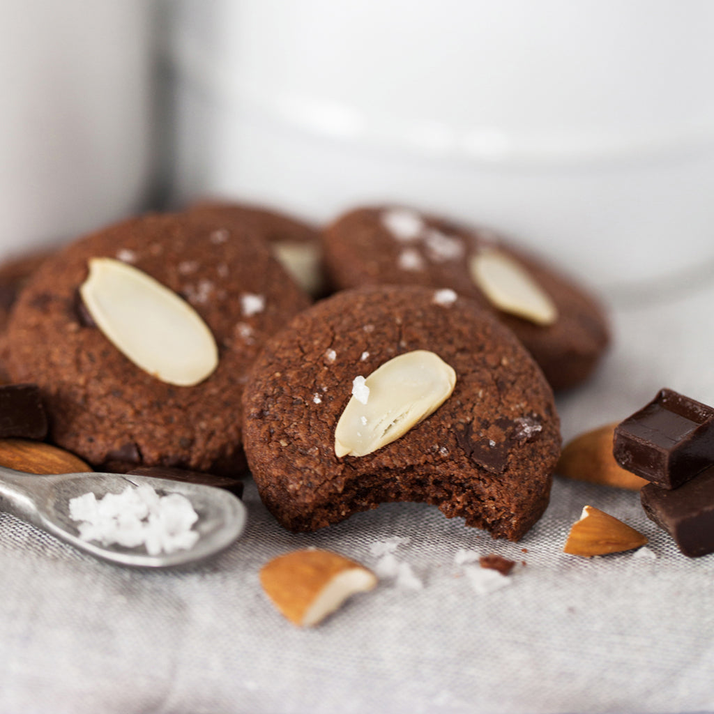 4 gluten-free Real Treat Pantry Dark Chocolate Almond with Sea Salt cookies sit on a table next to a smattering of sea salt, almonds and dark chocolate. 