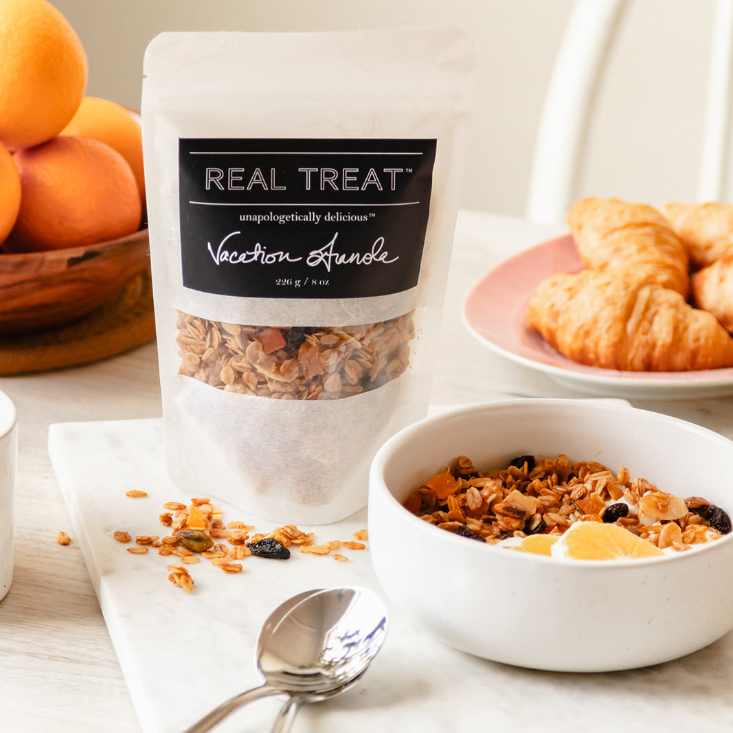 A package of Real Treat Vacation Granola is on a table next to a bowl of fruit and a bowl of granola with yogurt
