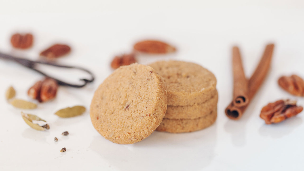 A stack of Real Treat Spiced Pecan Shortbread rests next to some scattered spices, including cardamom, cinnamon, vanilla beans and some pecans.