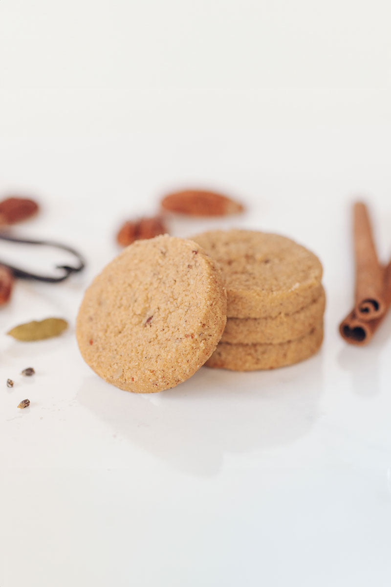 A stack of Real Treat Spiced Pecan Shortbread rests next to some scattered spices, including cardamom, cinnamon, vanilla beans and some pecans.