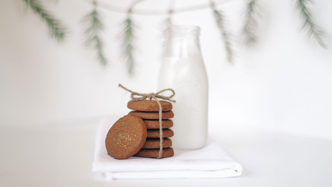 A stack of Real Treat Ginger Cookies next to a jug of milk, with a holiday garland in the background. 