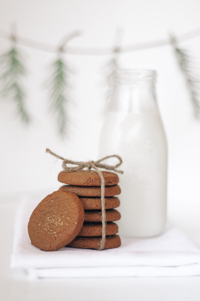 A stack of Real Treat Ginger Cookies next to a jug of milk, with a holiday garland in the background. 