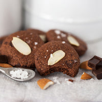 4 gluten-free Real Treat Pantry Dark Chocolate Almond with Sea Salt cookies sit on a table with a smattering of almonds, chunky sea salt, and dark chocolate.