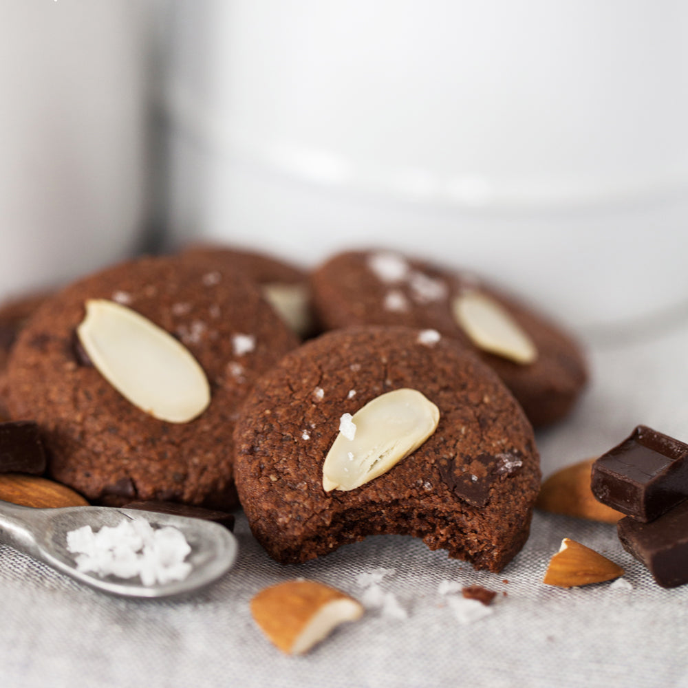 4 gluten-free Real Treat Pantry Dark Chocolate Almond with Sea Salt cookies sit on a table with a smattering of almonds, chunky sea salt, and dark chocolate. 