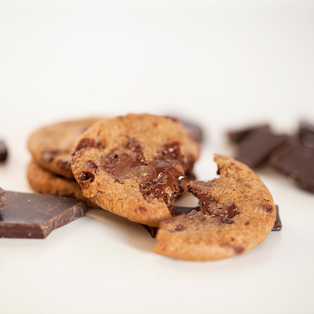 A single Real Treat Pantry Chocolate Chunk Cookie is broken in half with melted chocolate glistening. Next to the cookie are chunks of dark chocolate. 